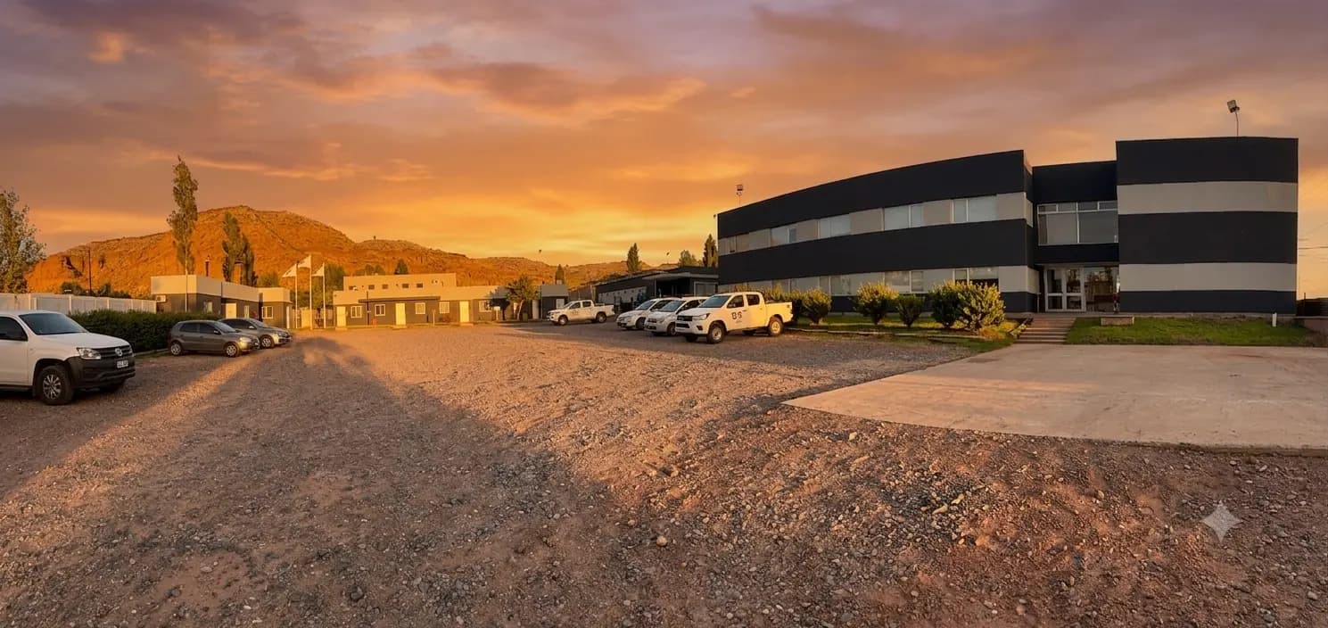 Fachada y estacionamiento del hotel en Añelo al atardecer, con camionetas de empresa y la barda al fondo.